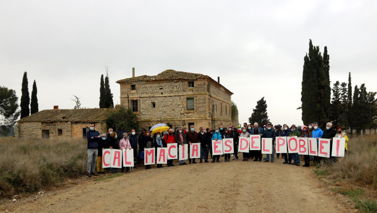 Pla general d'alguns dels participats a l'acte Vallmanya amb un cartell que reclama que "Cal Macià és del poble" i la casa al fons. Agència Catalana de Notícies